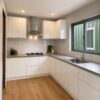 Kitchen interior of SAMAN affordable container home — white flat-panel cabinets, vinyl plank flooring, LED lighting, and aluminium-framed window