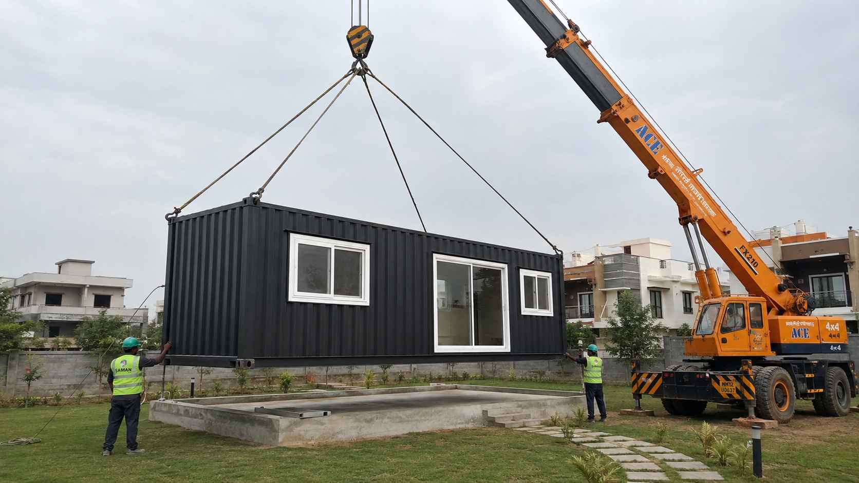A hydraulic crane lifting a SAMAN cargo container house into position above a prepared concrete plinth on a residential plot, with two installation crew in high-visibility vests guiding the placement.