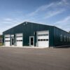 Teal commercial steel shed with white roller shutters, windows and an entry door on a commercial lot.