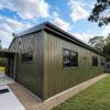 Olive-green prefabricated building side view with corrugated steel walls and black-framed windows.