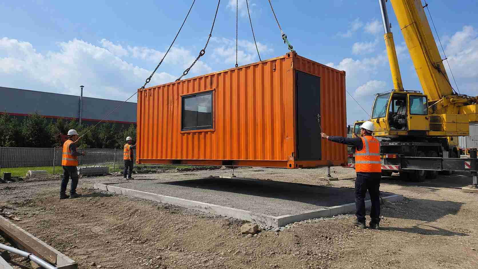 Orange container building lifted by crane onto a compacted base at a site.