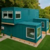 Modern blue container house exterior with large windows, patio seating, and bright landscaped surroundings.