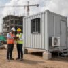 Two SAMAN site engineers in PPE reviewing project drawings beside a container portable office at an Indian construction site