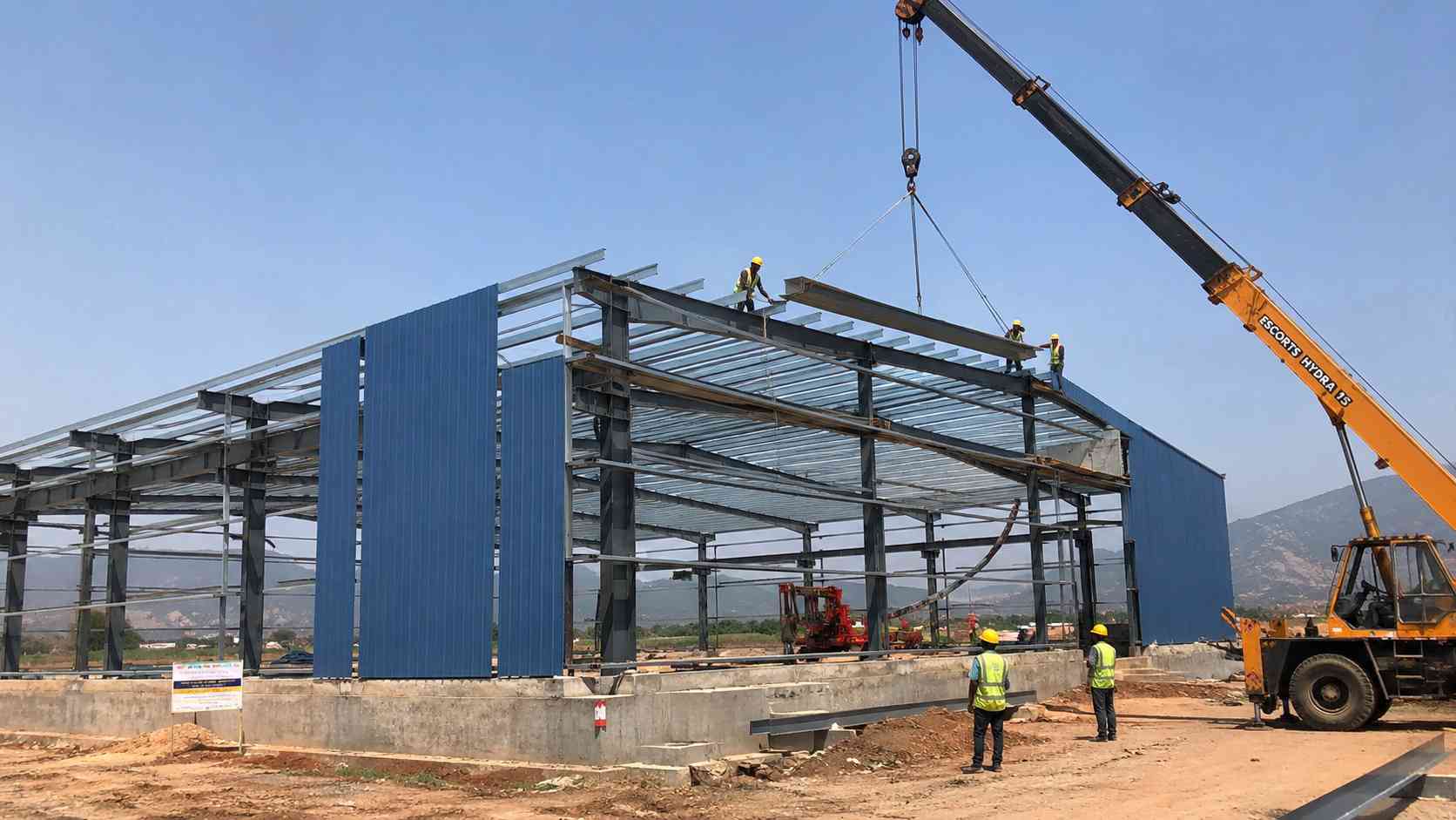 Steel-blue engineered building during erection, with a crane lifting a panel and crew on site.