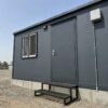A slate charcoal labor camp dormitory entry with steel door, brushed handle, welded step and concrete foundation block.