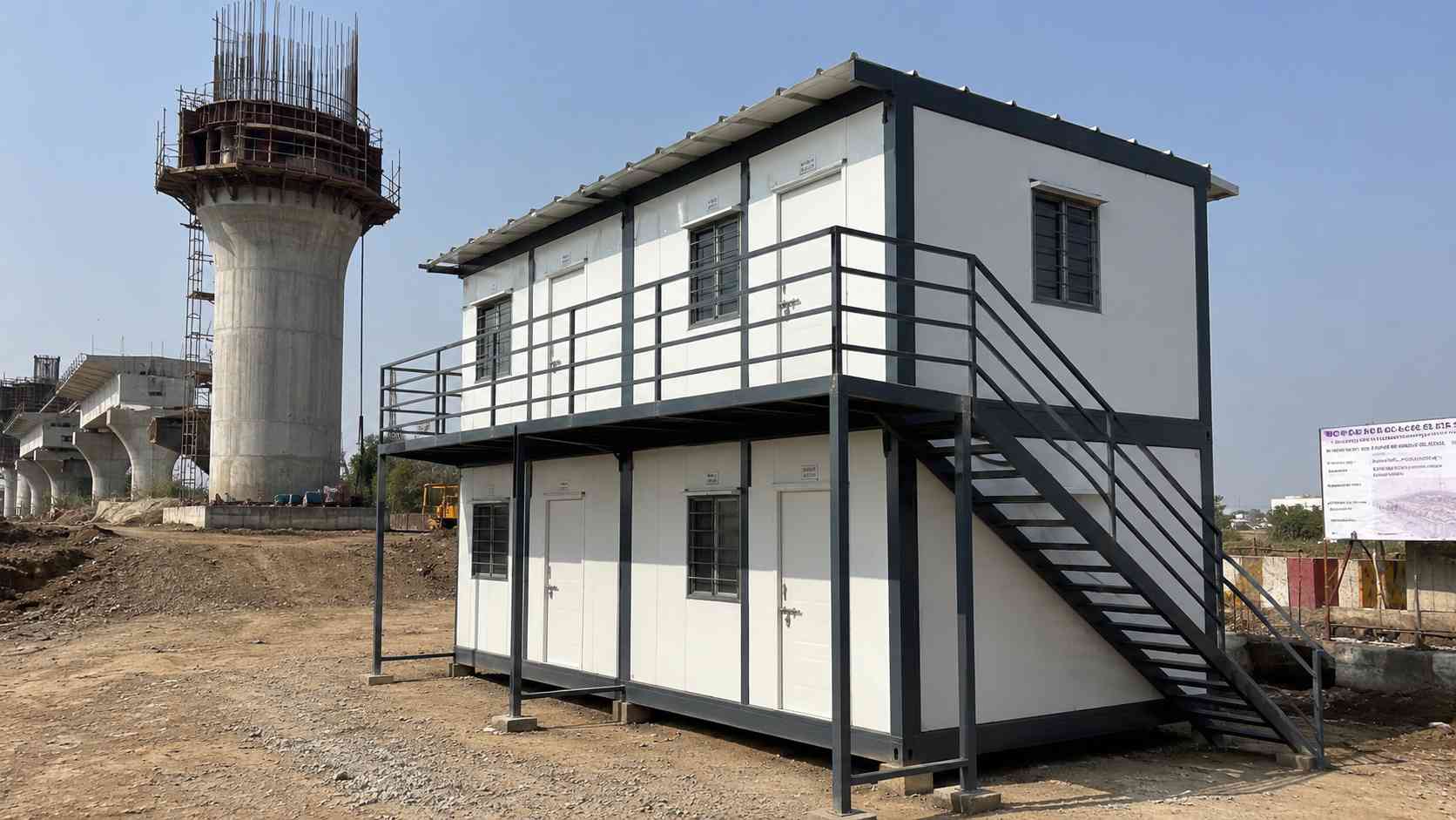 Two-tier SAMAN labor hutment block in Slate Charcoal beside a metro rail viaduct under construction.