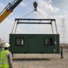 Forest green mobile container office mid-lift with spreader bar and four corner-casting slings at a transmission line site.