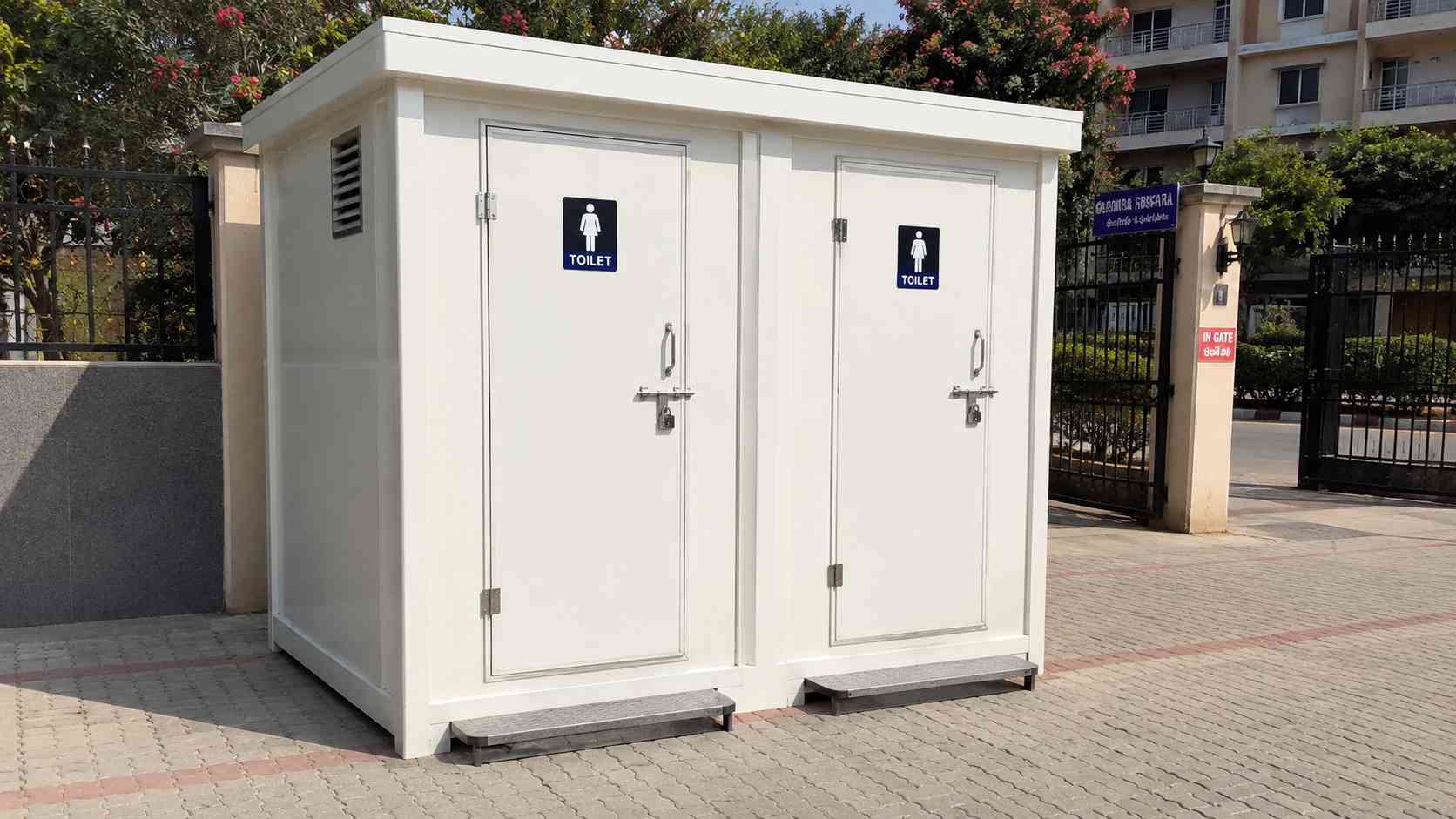 White two-cubicle mobile toilet cabin placed at a residential society compound entrance.