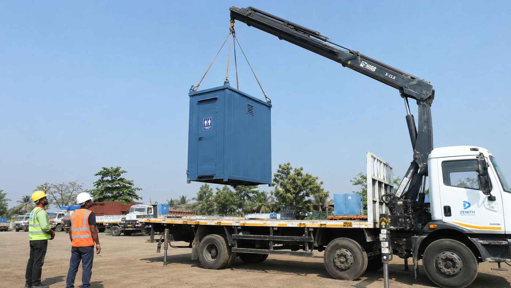 Steel blue mobile toilet cabin being safely crane-lifted onto a flatbed truck for relocation.