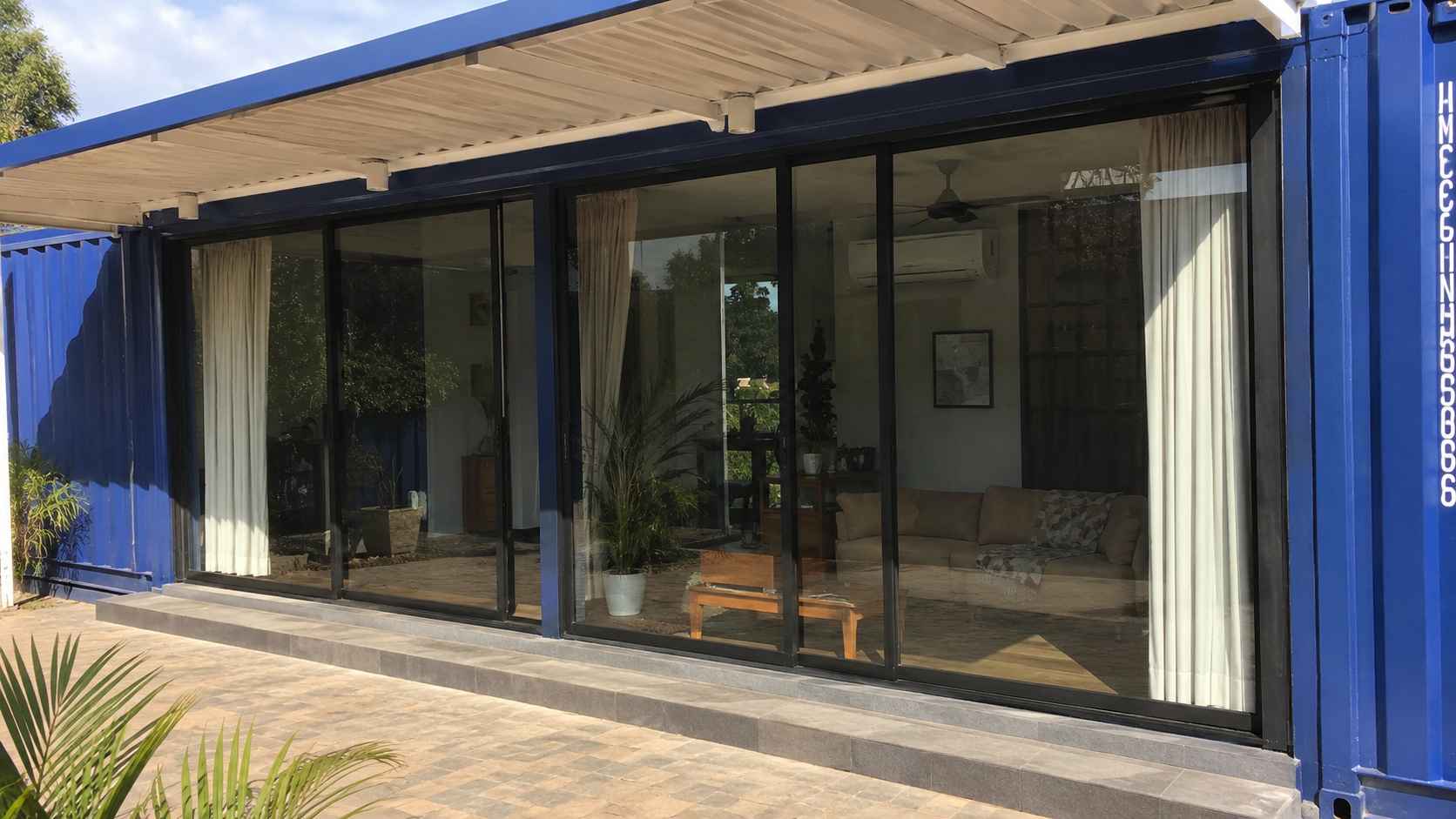 Large glazed windows shaded by a covered deck on a blue modern container home in sunlight.