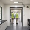 Engineers in PPE walking through corridor between two connected Forest Green modular container office units in active use.