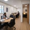 Interior of a modular portable office cabin in three-zone Layout B with a meeting table, two staff working at desks, and the manager cabin visible through an internal door.