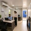 Office cabin interior in use, two professionals working at desks, false ceiling LED, partition, AC, MCB sockets visible.