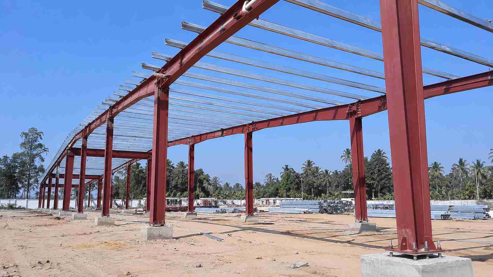 Bare red-oxide primer steel portal frames of a PEB being erected, purlins fixed across the top.