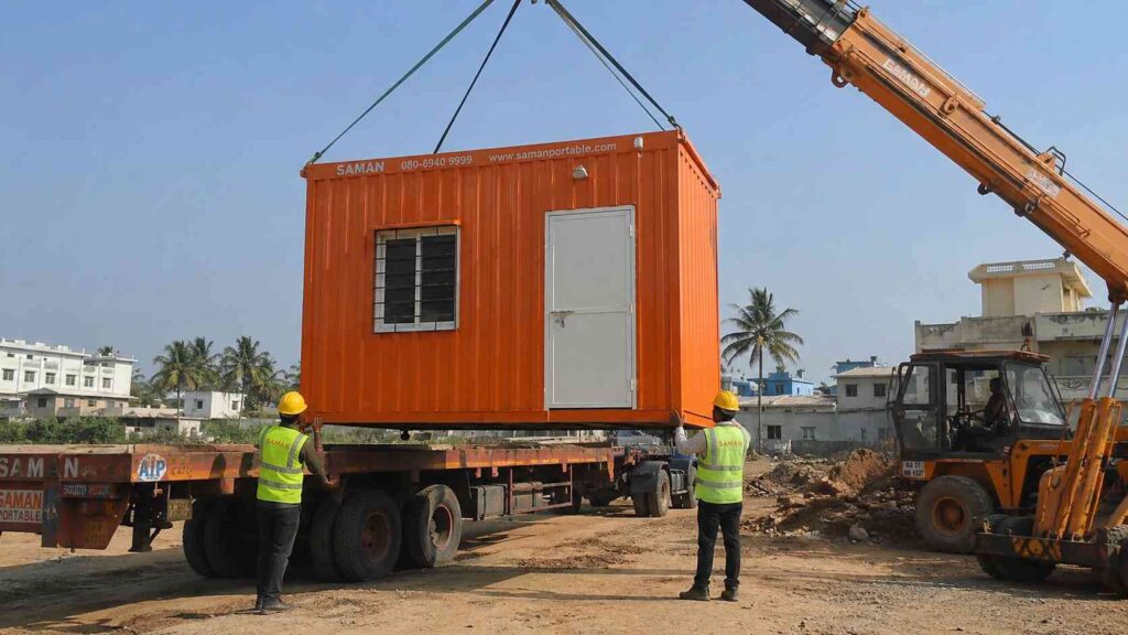 Safety orange porta cabin being unloaded from a 40 ft trailer at a Bangalore-area construction site.