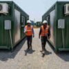 Two engineers in PPE walking between Forest Green portable container offices carrying drawings and project files.