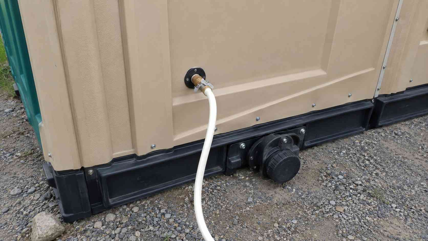 Water inlet and septic tank connection at the base of a beige portable toilet cabin