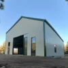 White pre-engineered building with green trim, an open vehicle bay and tall green-framed windows beside pine trees.