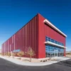 Red pre-engineered steel building with a glazed corner office entrance on a paved approach.
