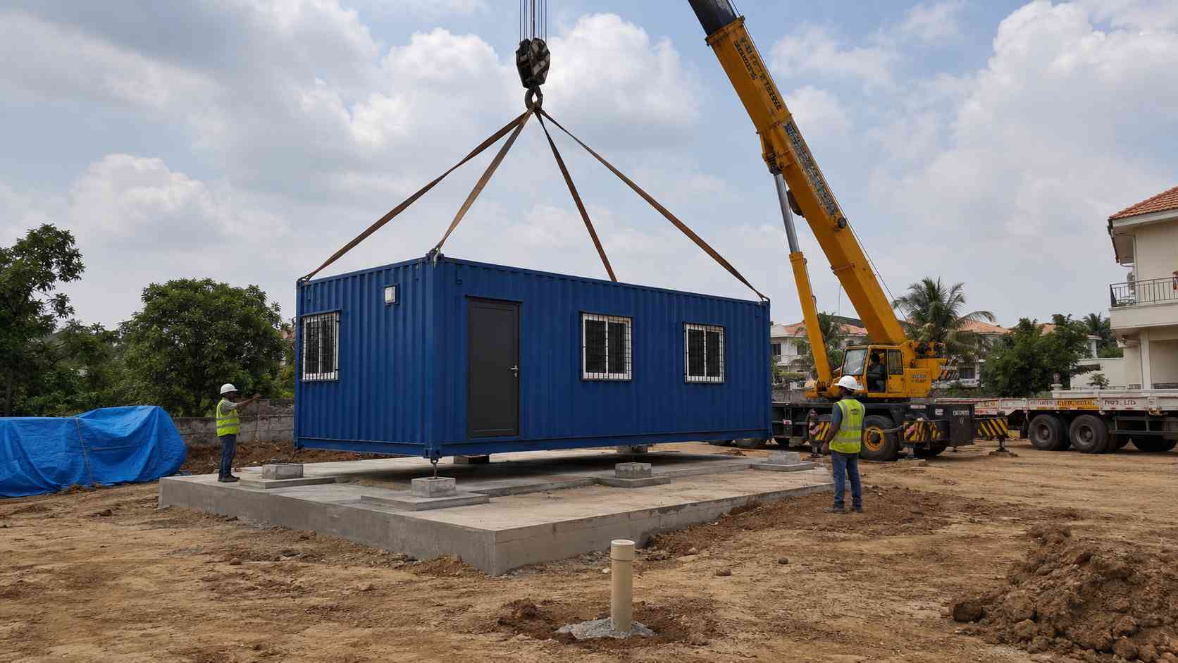 Mobile crane lowering a SAMAN Royal Cobalt prefab container home onto a prepared concrete plinth on an Indian residential site.