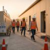 Construction workers in safety gear walk between dormitory and sanitation blocks at a SAMAN prefab labor camp