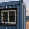 Macro of a Steel Blue prefab labor hutment window with black MS safety grille and edge-sealed panel joint.