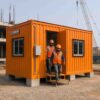 Workers in PPE walking out of a Safety Orange prefab labor hutment on an Indian project site.