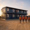 Workers in PPE returning to a navy blue G+1 prefab labor shed at the end of a shift on an Indian construction site