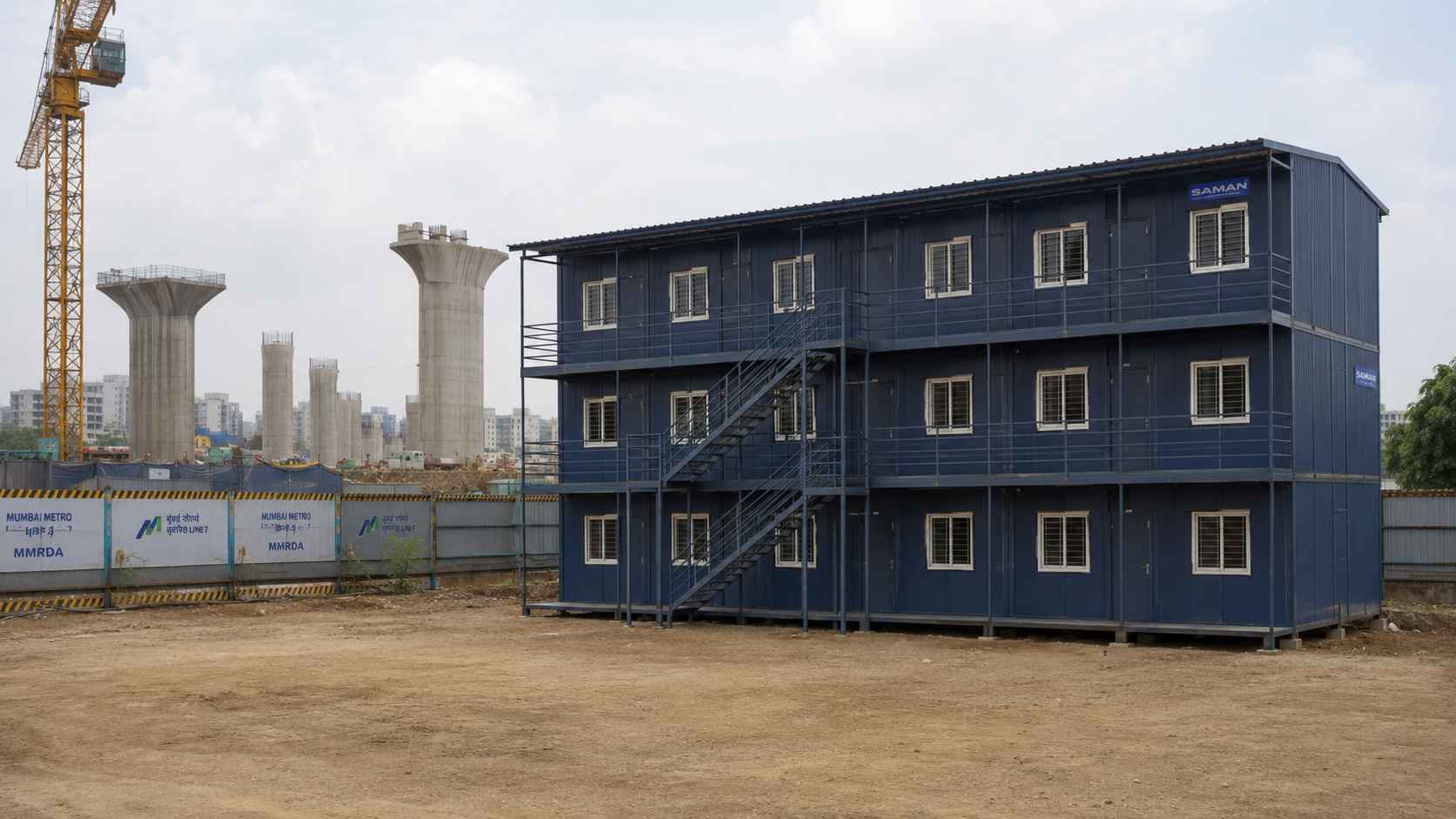 Navy blue G+2 prefab labor shed at a metro rail construction site on an Indian city outskirts with viaduct columns and tower crane in the background