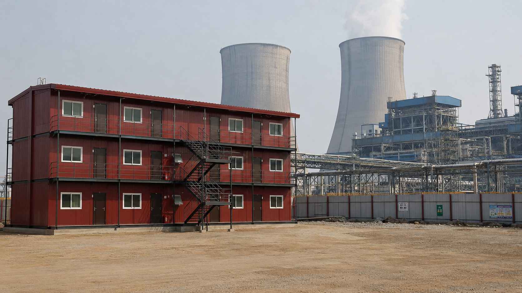 Burgundy red G+2 prefab labor shed at an Indian thermal power plant construction project with cooling towers and boiler-house structures in the background