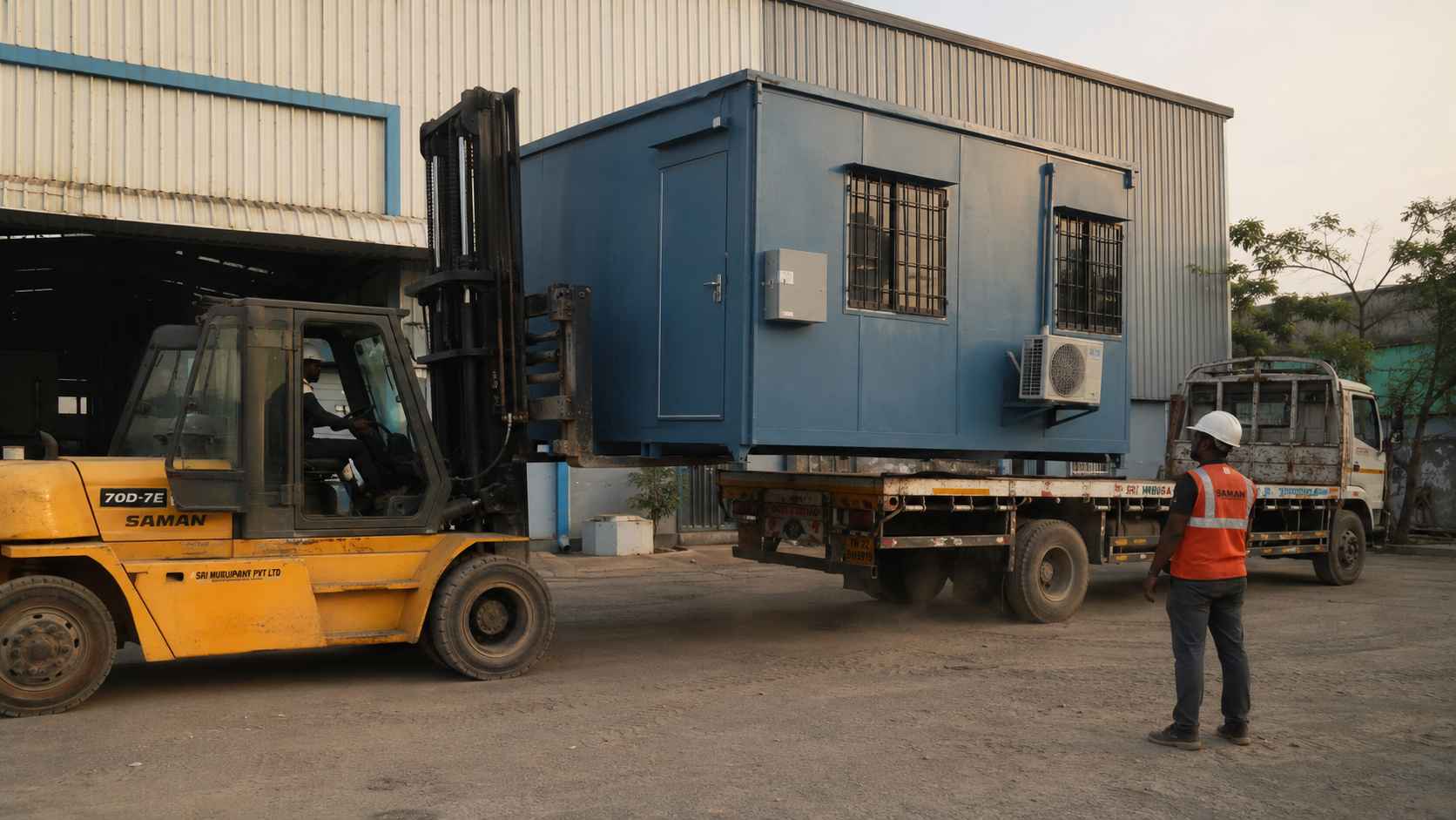 Steel Blue SAMAN cabin being loaded onto a flatbed by an industrial-yellow forklift outside an Indian warehouse.