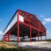 Red steel portal frame of a prefab modular construction building, partly clad, sides open during erection.