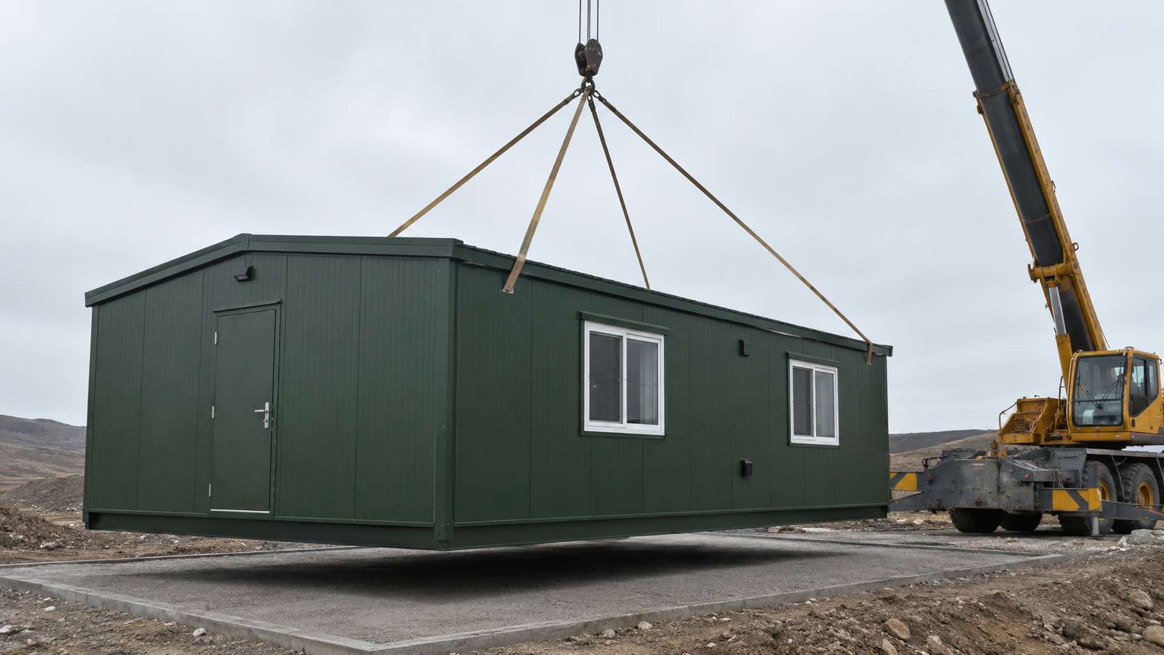 A forest green prefabricated bunkhouse being craned onto a level base at a remote project site.