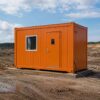 A safety-orange prefabricated bunkhouse connected to power at the edge of a worksite.
