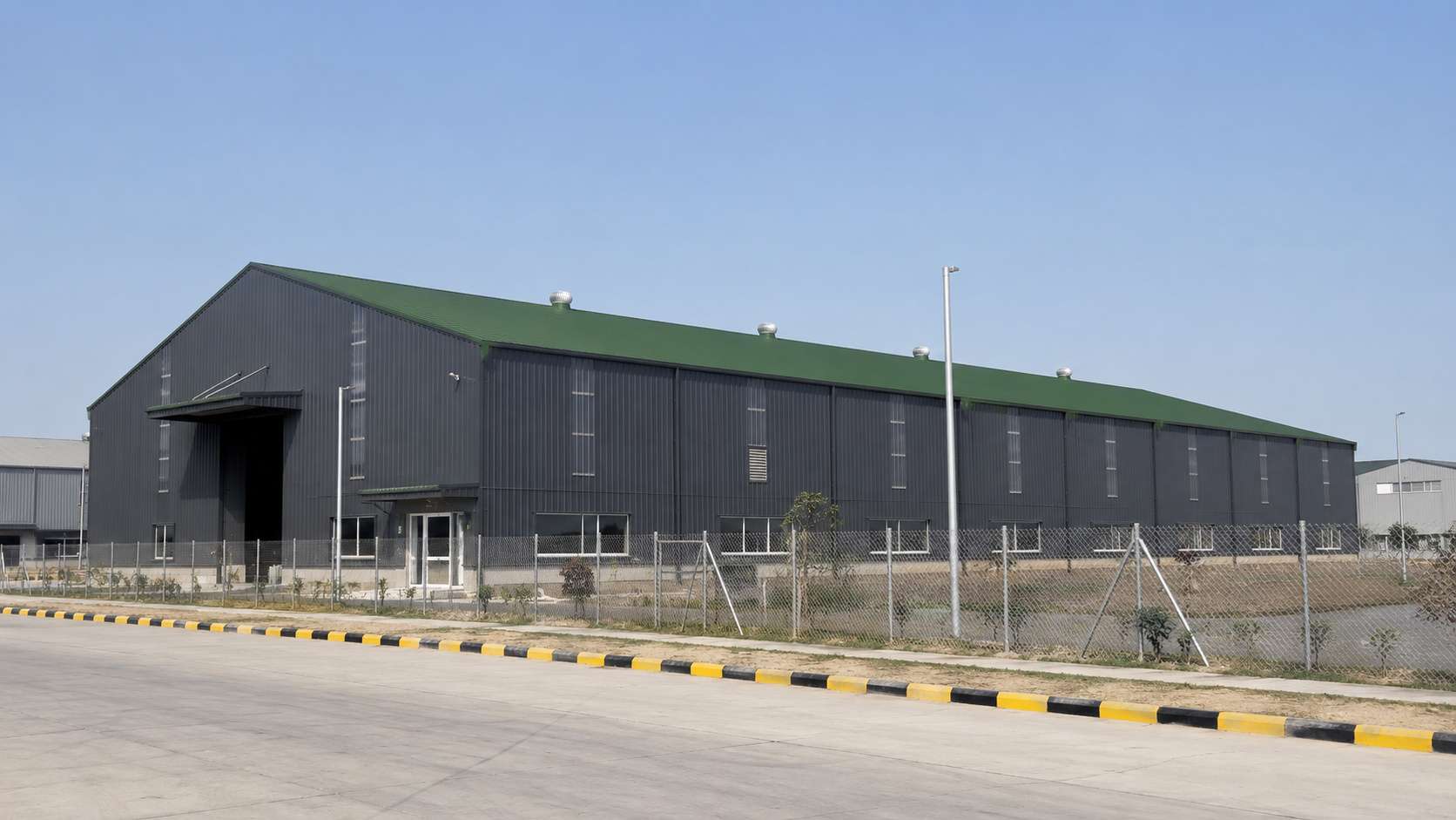 Charcoal-grey prefabricated steel building with a green steel roof on an industrial estate road.