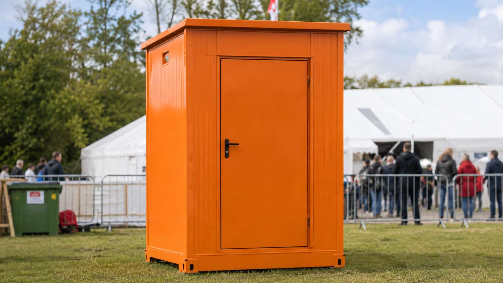 Safety orange readymade toilet cabin at an outdoor event ground with a marquee behind.