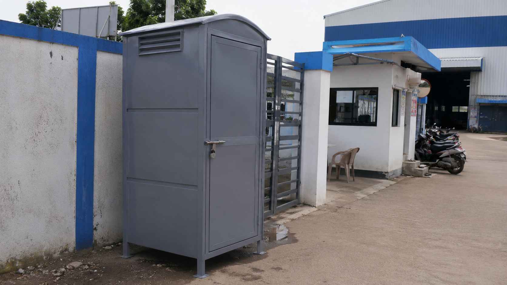 Steel grey readymade toilet cabin placed beside a factory gate and security point.