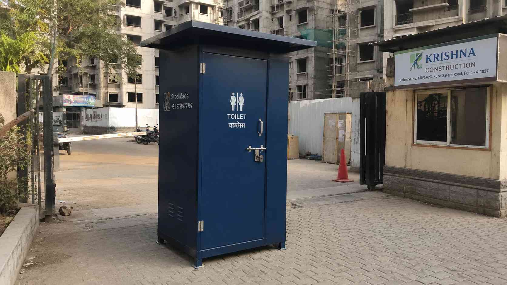Navy blue readymade toilet cabin at a residential society construction entrance.