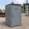 Steel grey readymade toilet cabin on a gravel pad at a construction site.