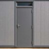 Front entrance door detail of prefab building with steel frame, solid grey panels, transom window, and factory-precision joints