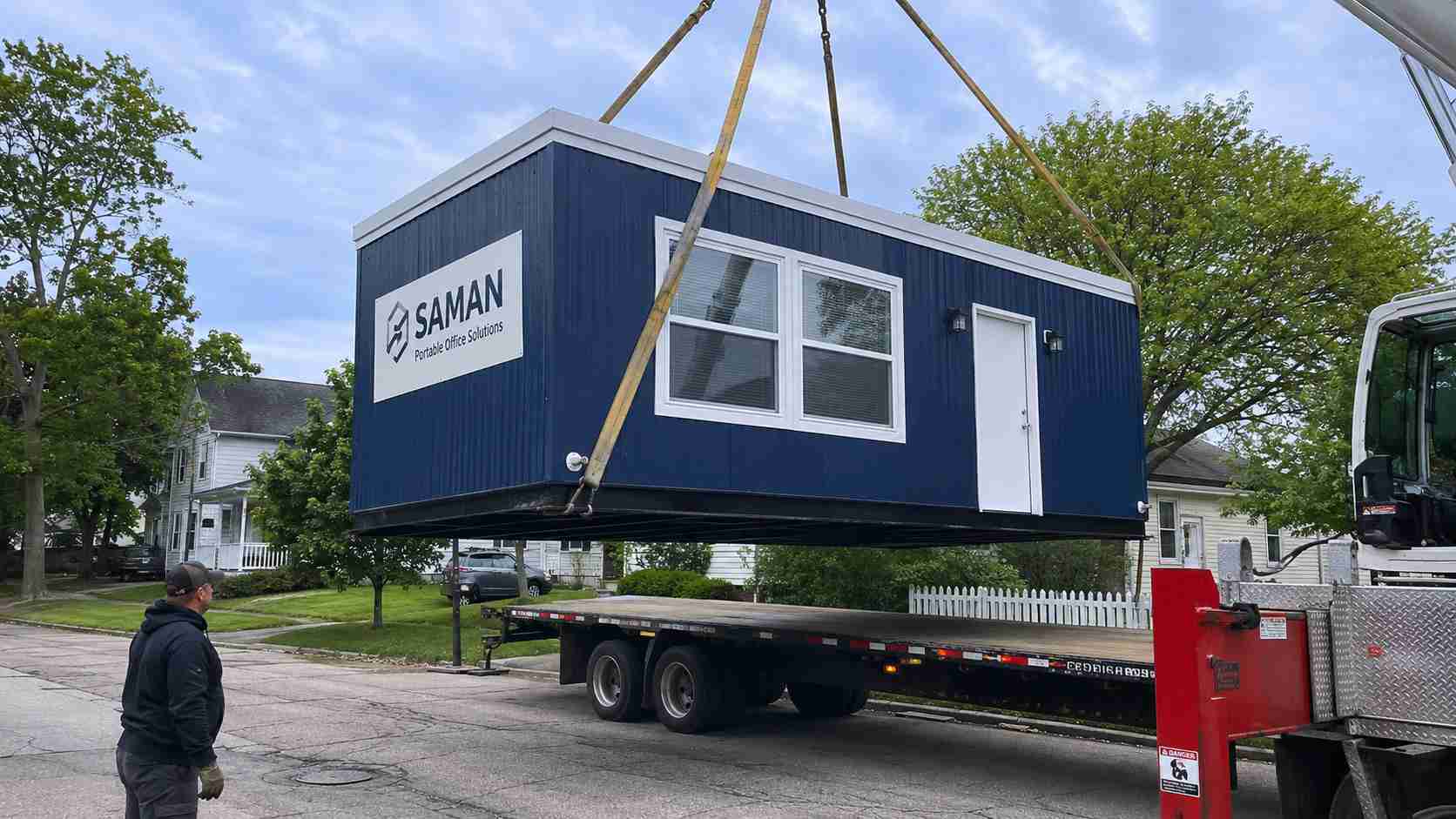 Navy blue SAMAN prefab office cabin loaded onto a flatbed for relocation in a residential locality.