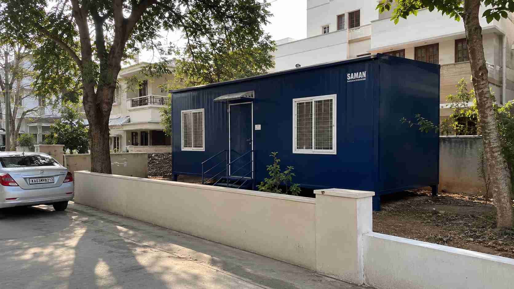 Navy blue SAMAN prefab office on an owned residential plot behind a boundary wall, quiet street.