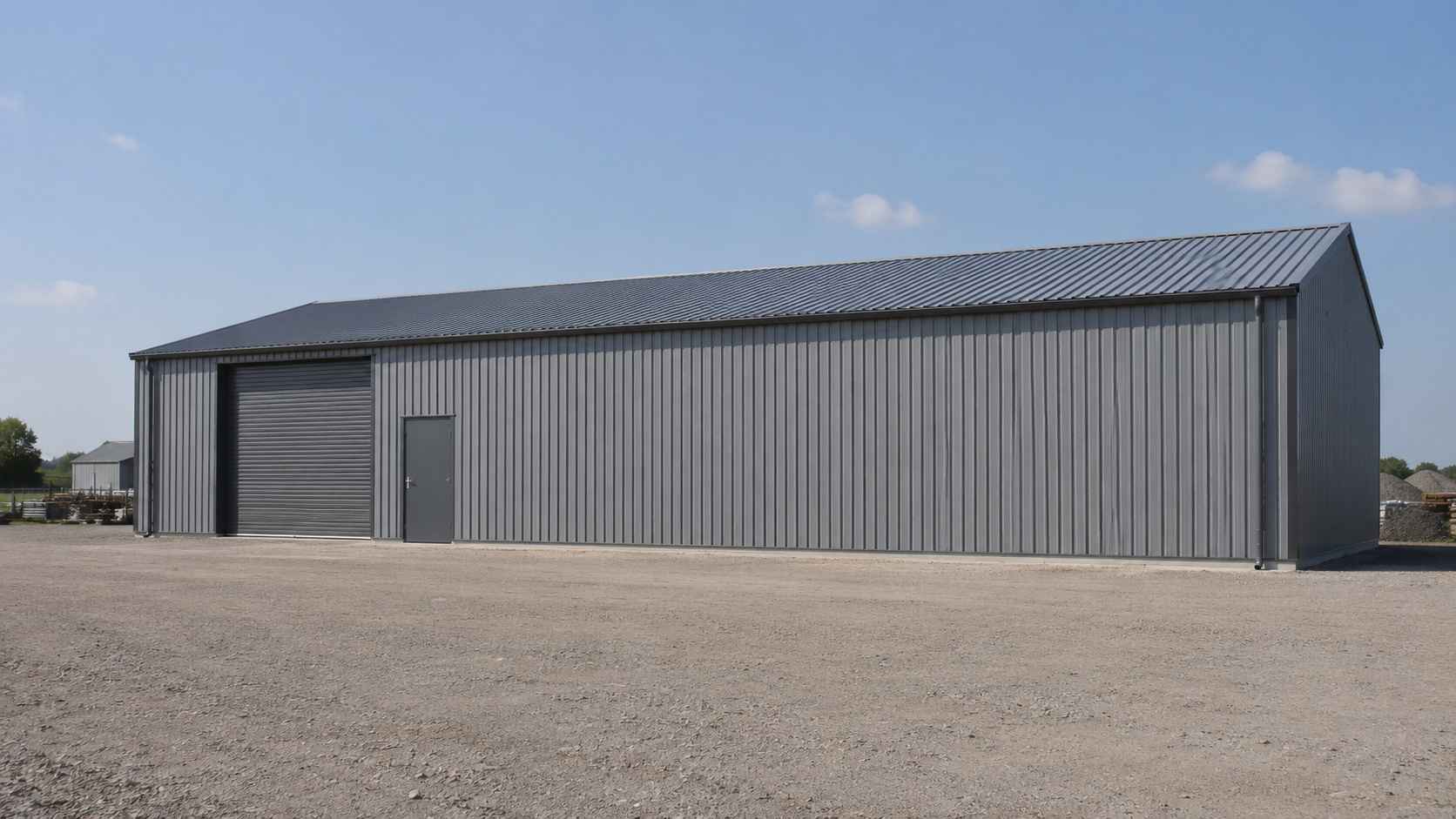 A long galvanised steel-grey shed on an industrial plot, showing the full pitched-roof side elevation