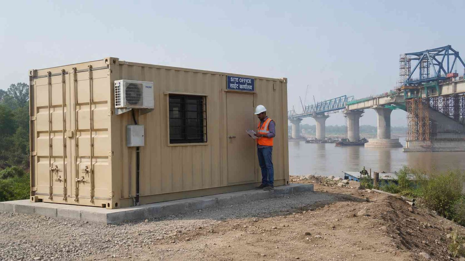 Sandstone Beige SAMAN storage container office at an Indian river bridge construction site with a supervisor at the office door and the partially-completed bridge visible in the background