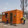 Two Indian engineers in PPE working at a Safety Orange SAMAN storage container office at the base of a telecom BTS tower with the office door open showing the internal partition