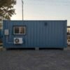 Side elevation of a SAMAN Steel Blue 20-ft storage container office showing split AC, electrical distribution box, sliding window on the office side and a storage-side ventilation grille