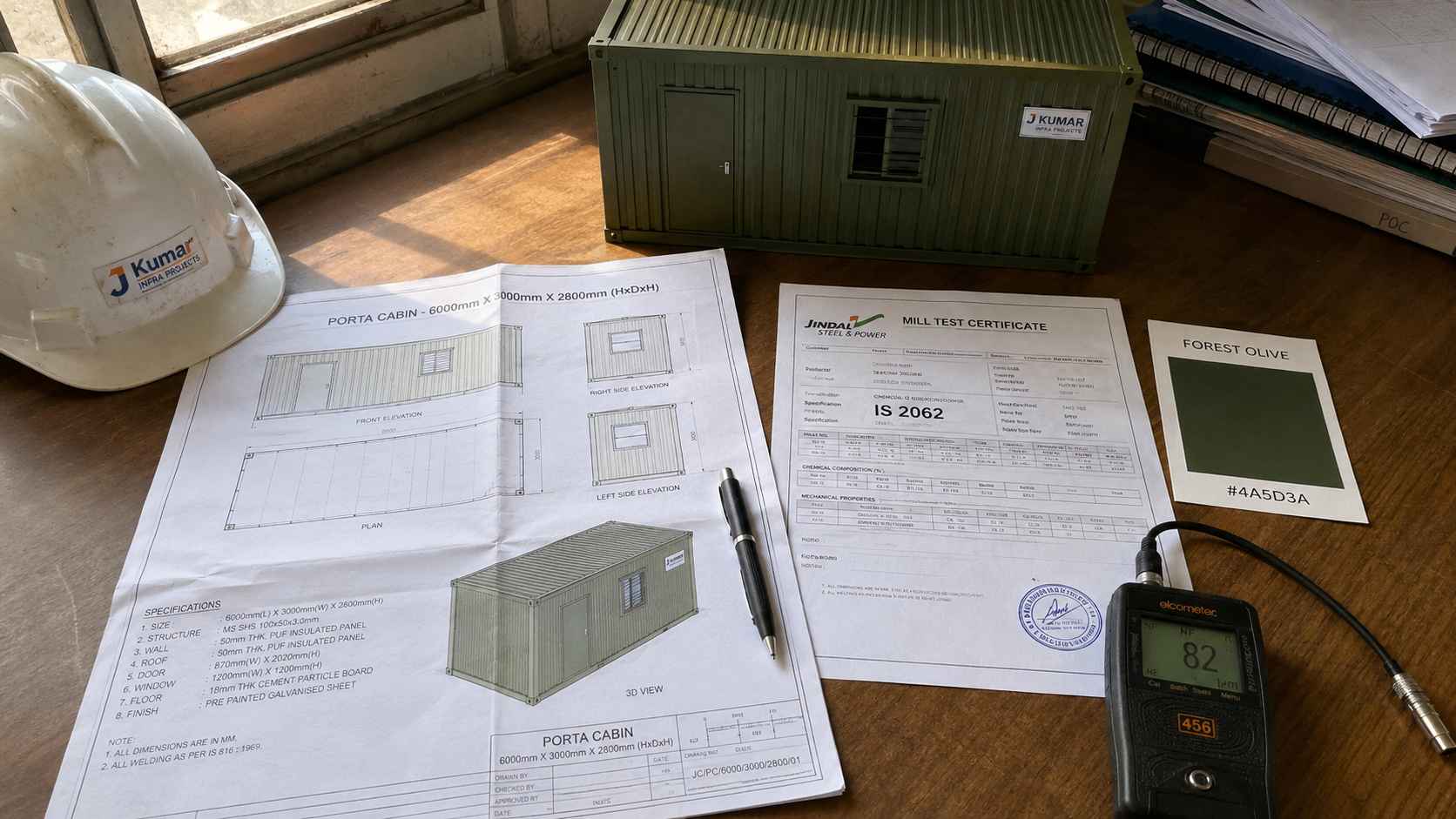 Porta cabin procurement documentation flat-lay — mill test certificate, structural drawing, DFT gauge, and Forest Olive colour scale model on an Indian project office desk.