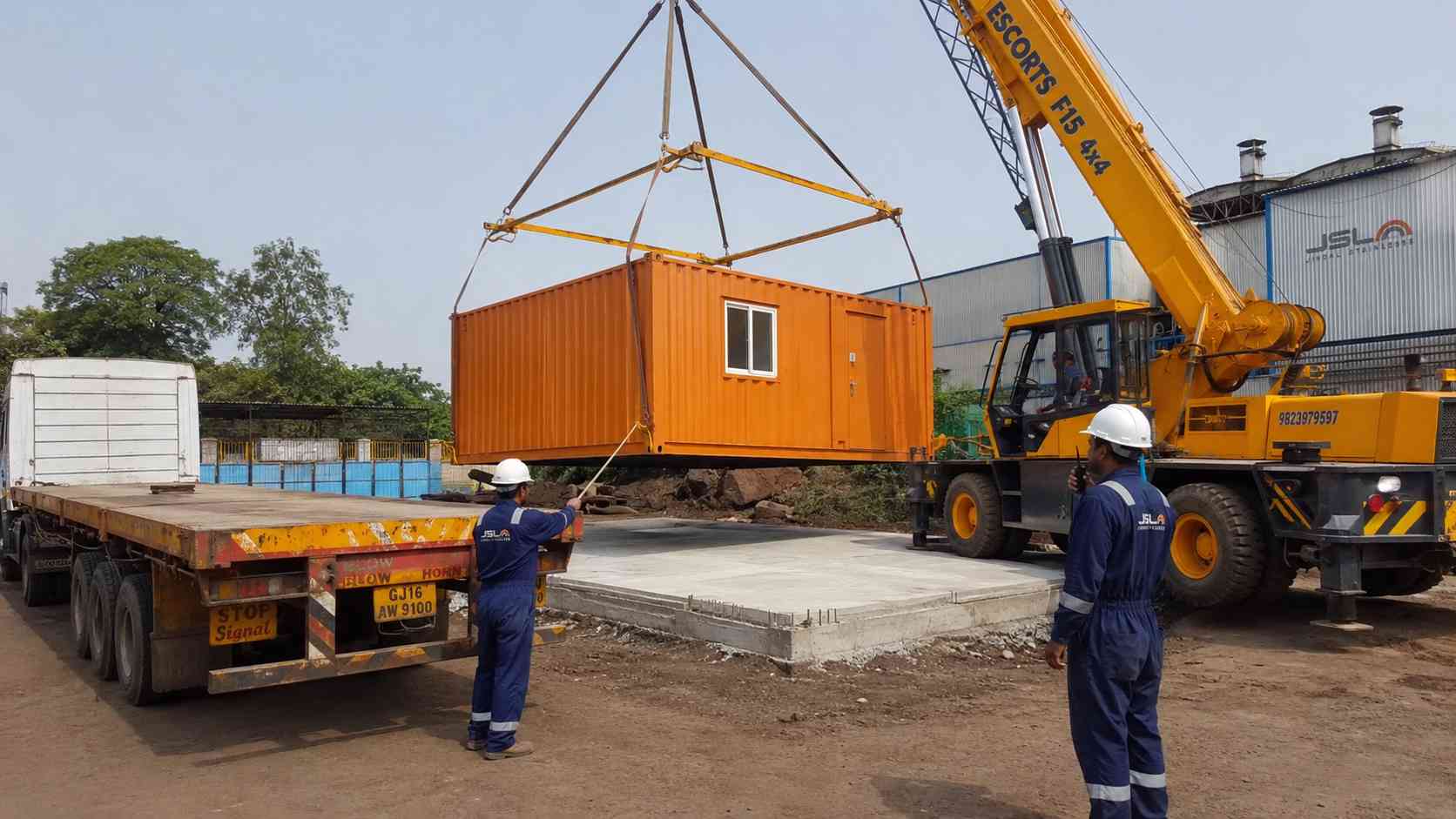 Hydraulic crane lifting a Safety Orange #D17A1F 20ft container office onto a foundation pad at an Indian industrial site, showing the delivery and installation costs.