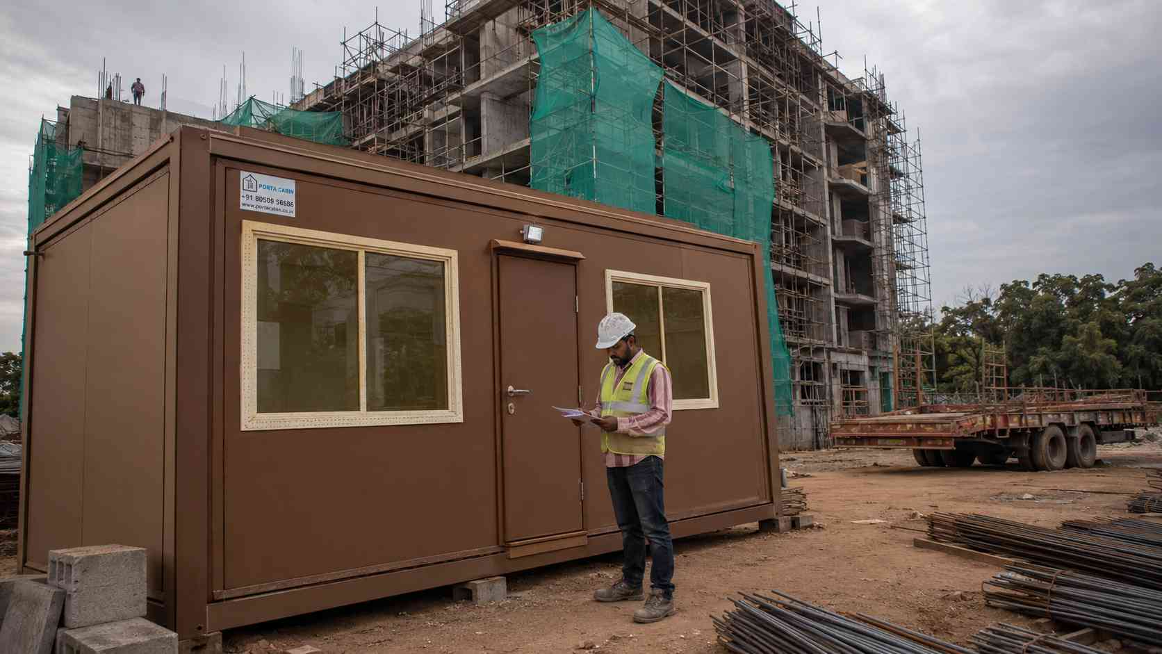 Earth Umber 20×10 ft 2-room porta cabin functioning as an apartment-build site office on a south-Bangalore redevelopment site, with scaffolding rising in the background.