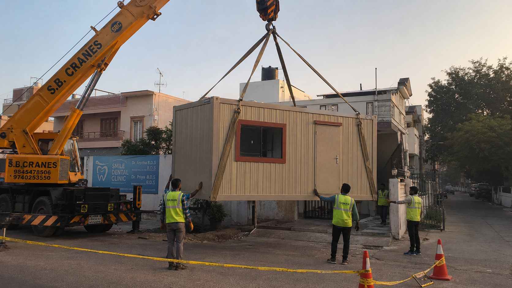 Quartz Beige 10×16 ft single-room porta cabin being hydra-crane lifted into placement on a BTM 1st Stage residential-conversion plot at early-morning installation, with the SAMAN crew coordinating the lift.